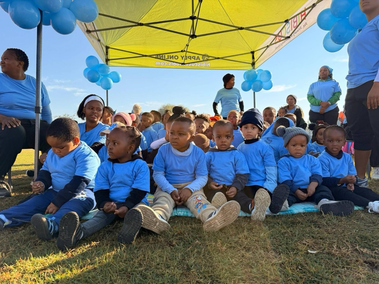 Toddlers in blue T-shirts sitting together under a Hibiscus gazebo at sports day